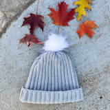 gray ribbed crochet hat with a white pom pom laying on the ground surrounded by colorful leaves