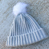 gray ribbed crochet hat with a white pom pom laying on the ground