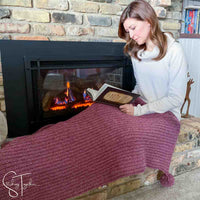 lady reading a book while draped in a red holiday crochet blanket next to a fireplace