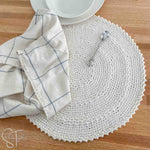 crochet placemat laying flat with a small spoon, dishtowel and some dishes on it
